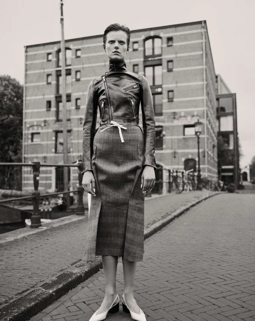 A woman in a leather jacket and skirt poses on a cobblestone street in front of a brick building in a black and white fashion photograph.