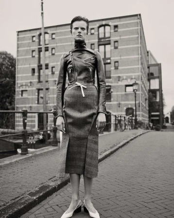 A woman in a leather jacket and skirt poses on a cobblestone street in front of a brick building in a black and white fashion photograph.
