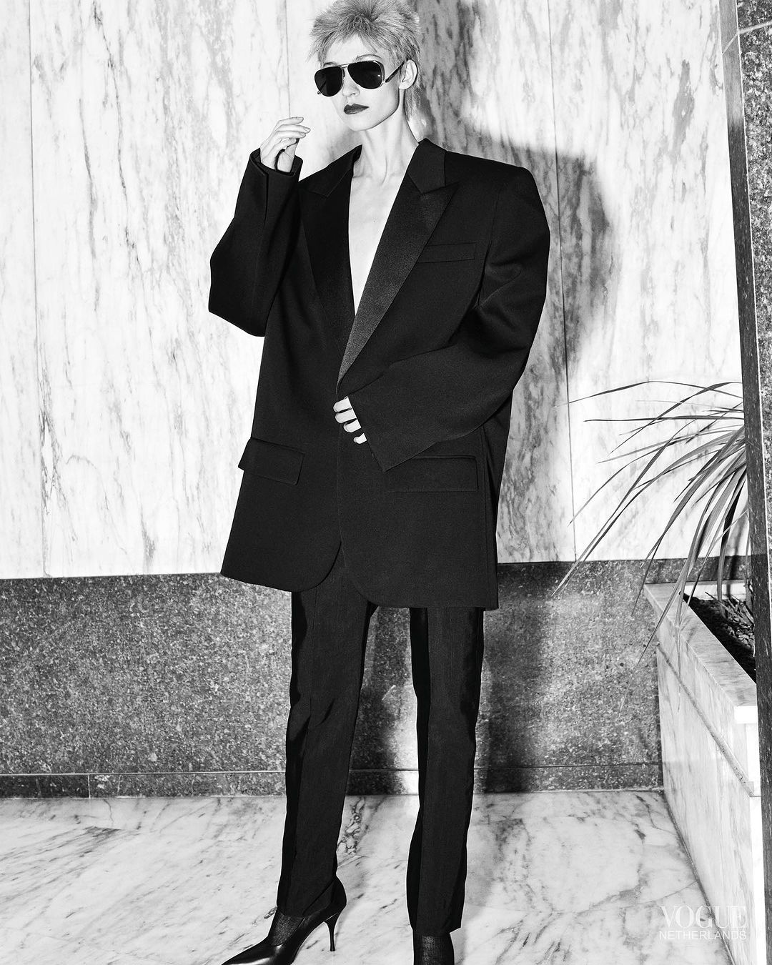 An adult woman with spiky blonde hair is posing in a black suit and heels in front of a marble wall, with sunglasses. Fashion editorial.