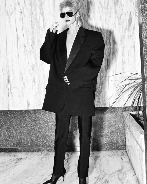 An adult woman with spiky blonde hair is posing in a black suit and heels in front of a marble wall, with sunglasses. Fashion editorial.