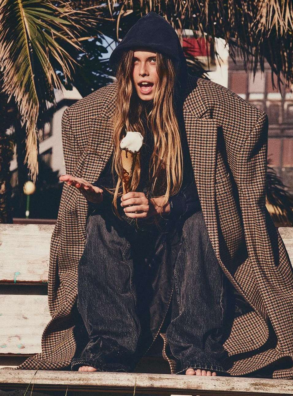 A person sits on a wooden bench outdoors wearing an oversized houndstooth coat and holding ice cream with palm trees in background.