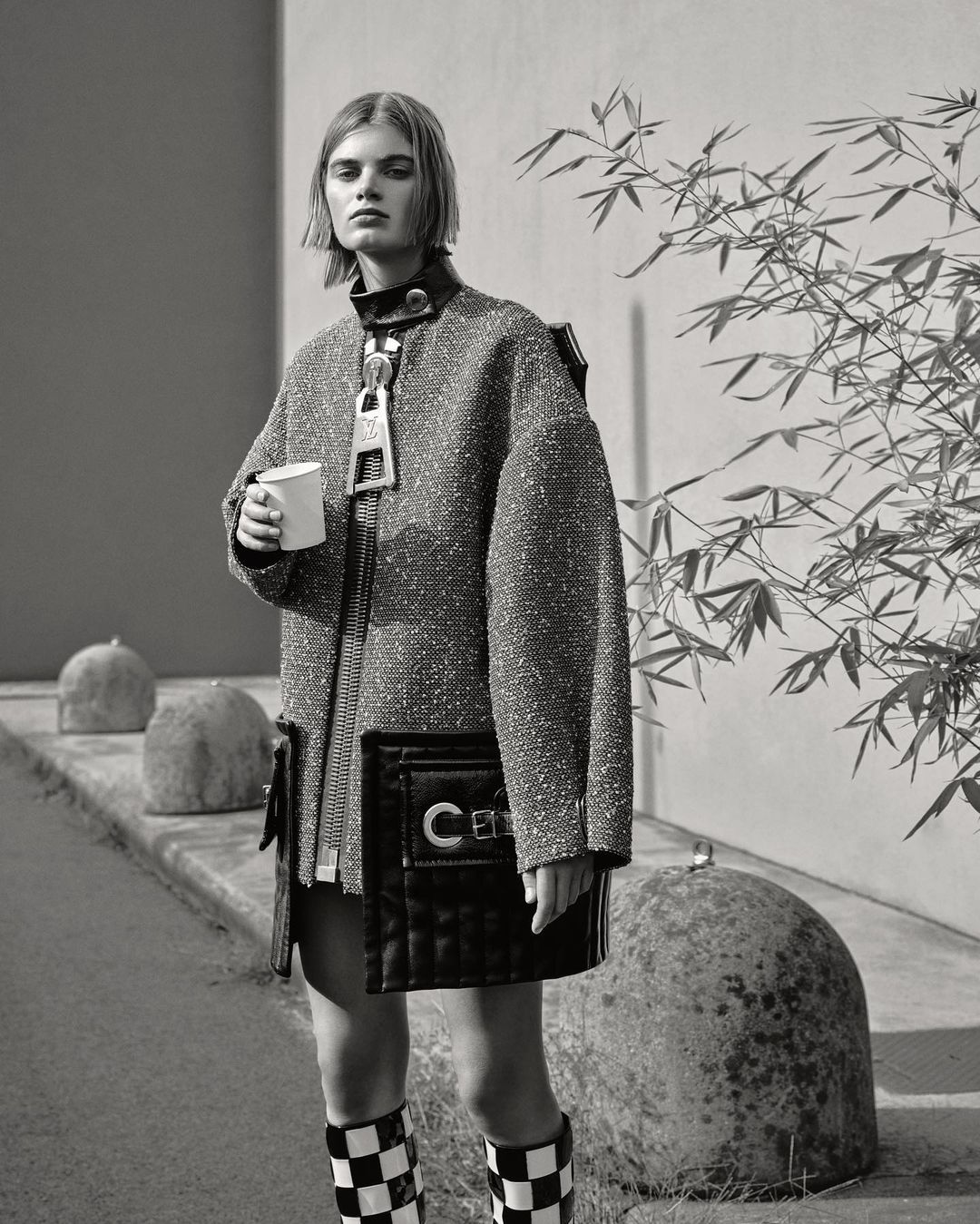 Fashion shot of a woman wearing a textured coat and checkered boots, holding a cup, with rocks and greenery in the background.