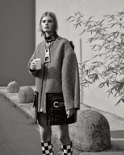 Fashion shot of a woman wearing a textured coat and checkered boots, holding a cup, with rocks and greenery in the background.