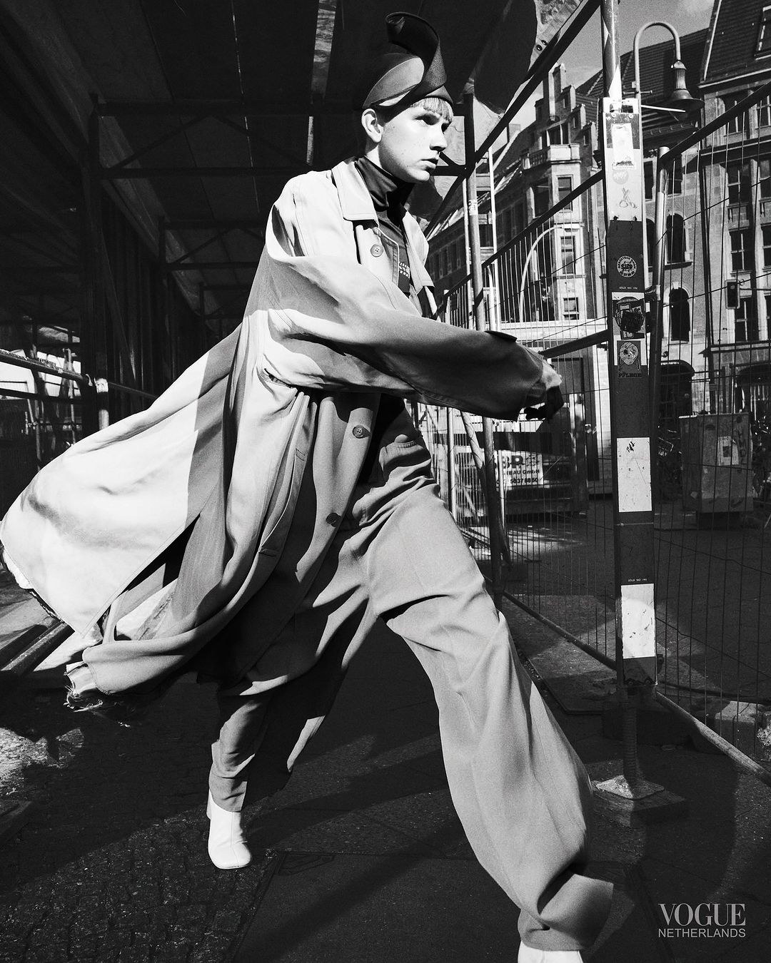 Black and white fashion shot of a young adult walking outdoors wearing an oversized trench coat, wide leg pants, and a pointed hat.