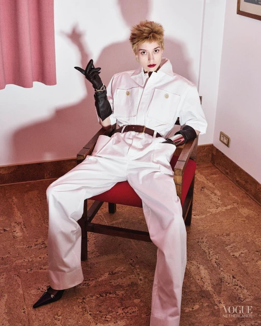 Stylish woman in a white jumpsuit and black gloves sits in a red chair indoors, posing with a raised hand, against a light pink wall.