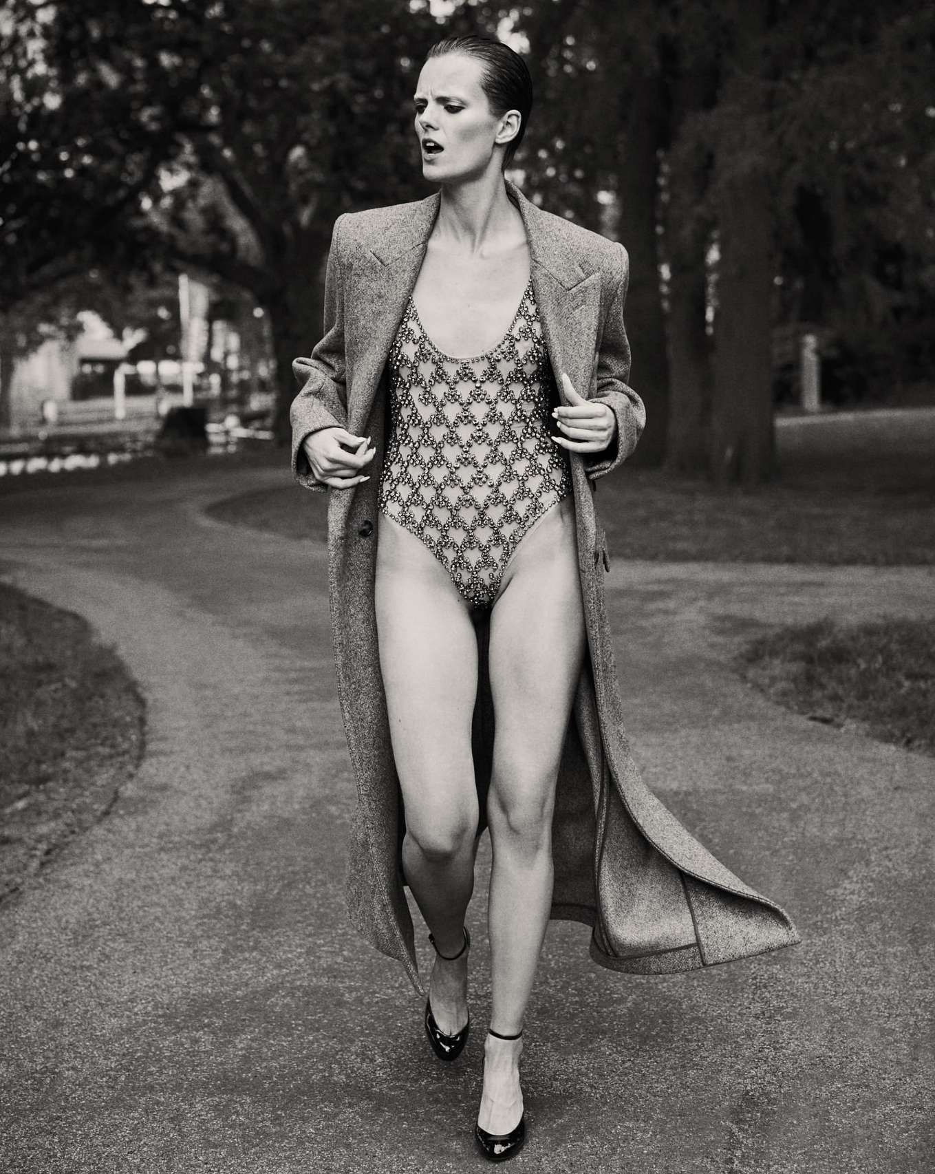 A model walks on a park path wearing a patterned swimsuit, a long gray wool coat, and high-heeled shoes in a black and white fashion shot.