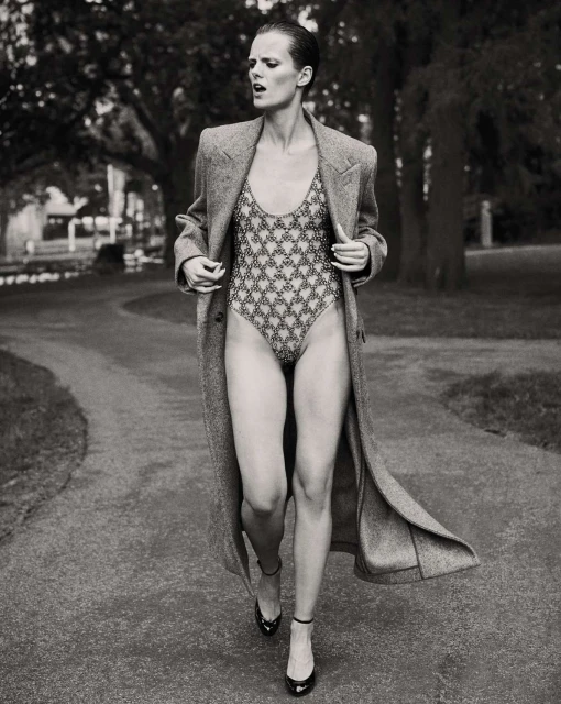 A model walks on a park path wearing a patterned swimsuit, a long gray wool coat, and high-heeled shoes in a black and white fashion shot.