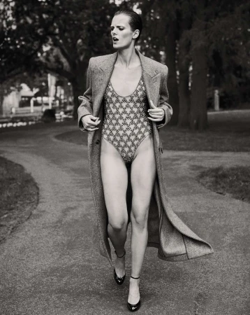A model walks on a park path wearing a patterned swimsuit, a long gray wool coat, and high-heeled shoes in a black and white fashion shot.