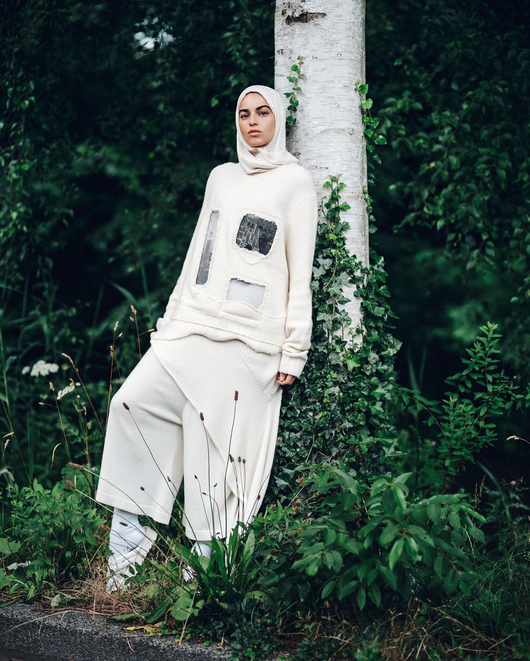 A woman in a headscarf and cream-colored outfit poses next to a birch tree in a green outdoor setting, blending fashion and nature.