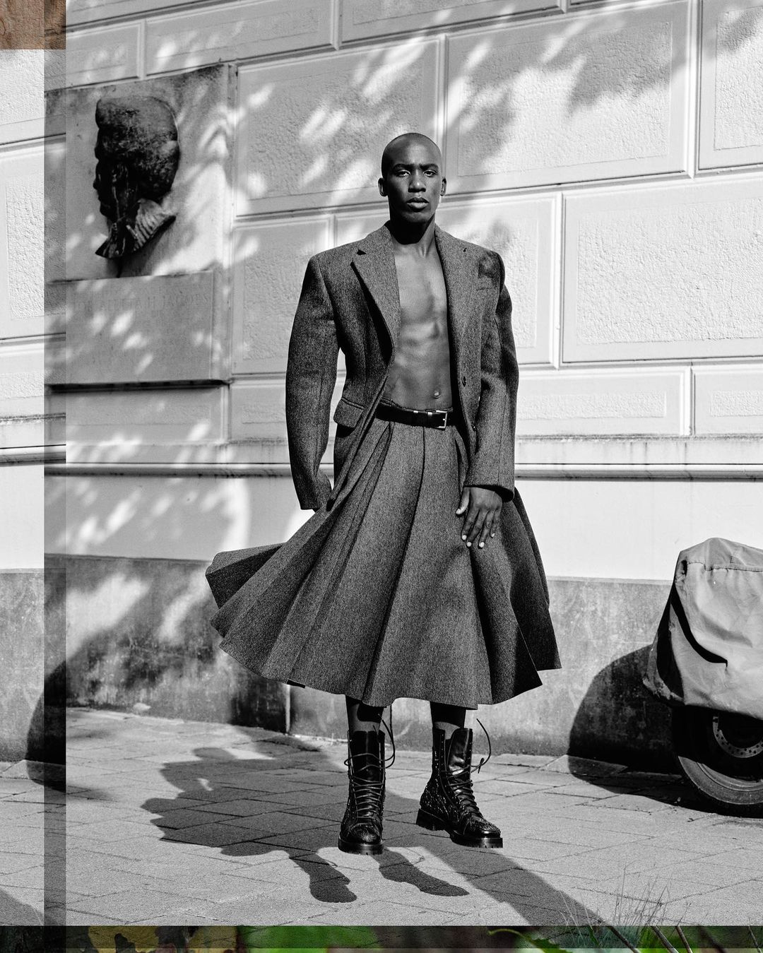 Monochrome full shot of a Black male fashion model wearing an open wool coat and pleated skirt with combat boots against a building.