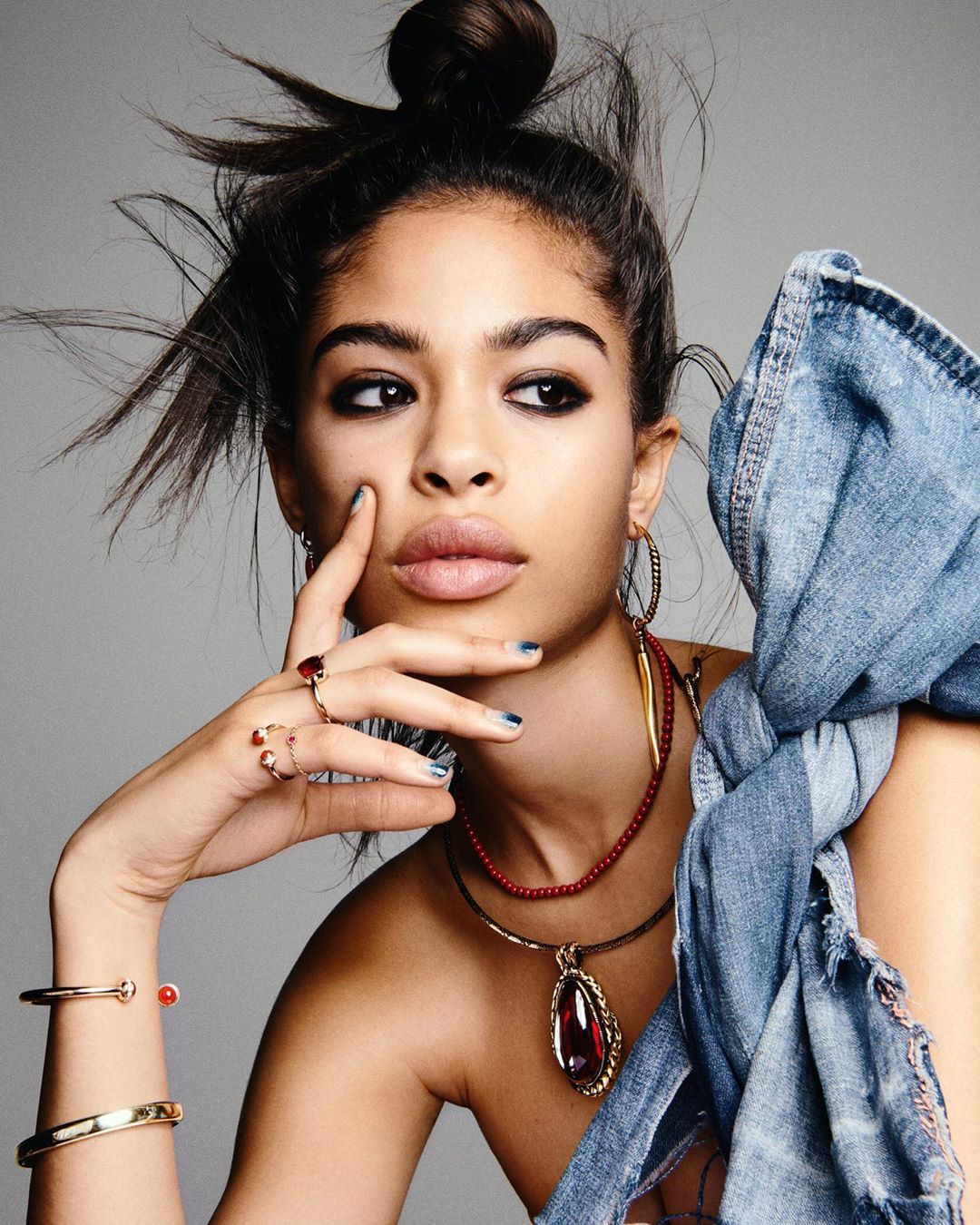 Close-up portrait of a young woman with dark hair in a high bun, wearing statement jewelry, against a neutral grey studio backdrop.