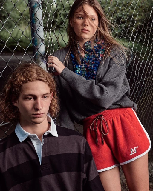 Two young adults pose by a chain-link fence; one wears a striped shirt, while the other is in red shorts and a gray cardigan.
