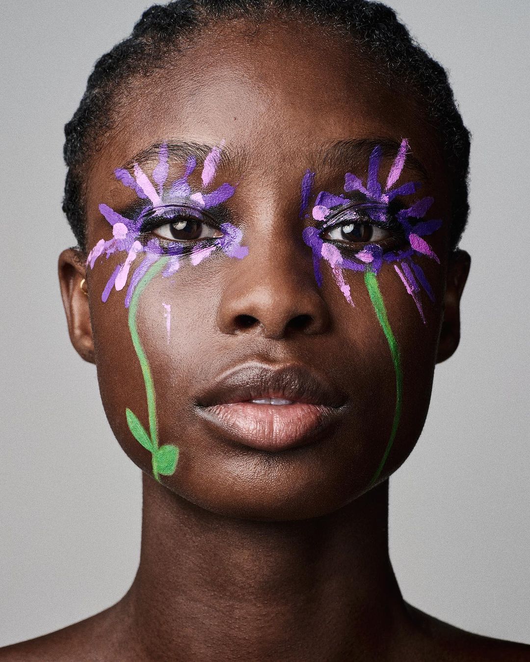 Close-up portrait of a young Black woman with colorful flower makeup around her eyes, set against a neutral grey studio backdrop.