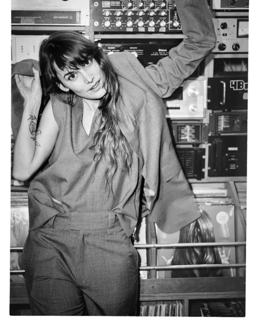 Black and white portrait of a woman in a suit, leaning against an audio soundboard with records on shelves in the background.