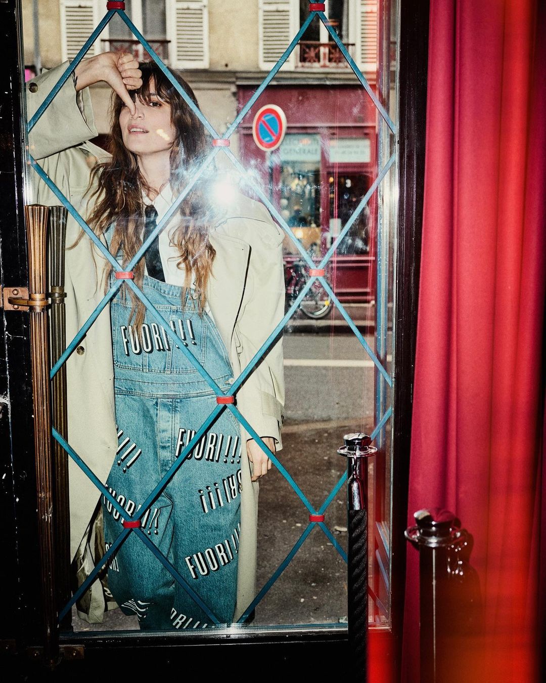 A stylish woman poses in a window wearing denim overalls with text. She is in an urban setting with a building in the background.