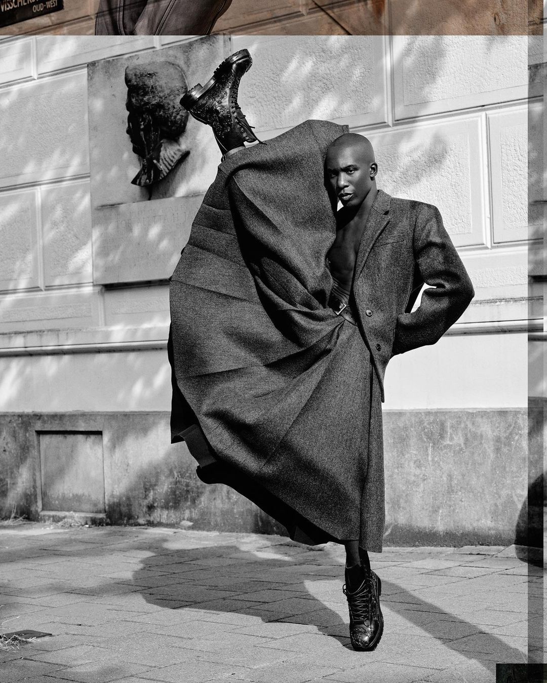 Black and white fashion shot of a Black male model in tweed coat and long skirt, balanced on one foot in front of building.