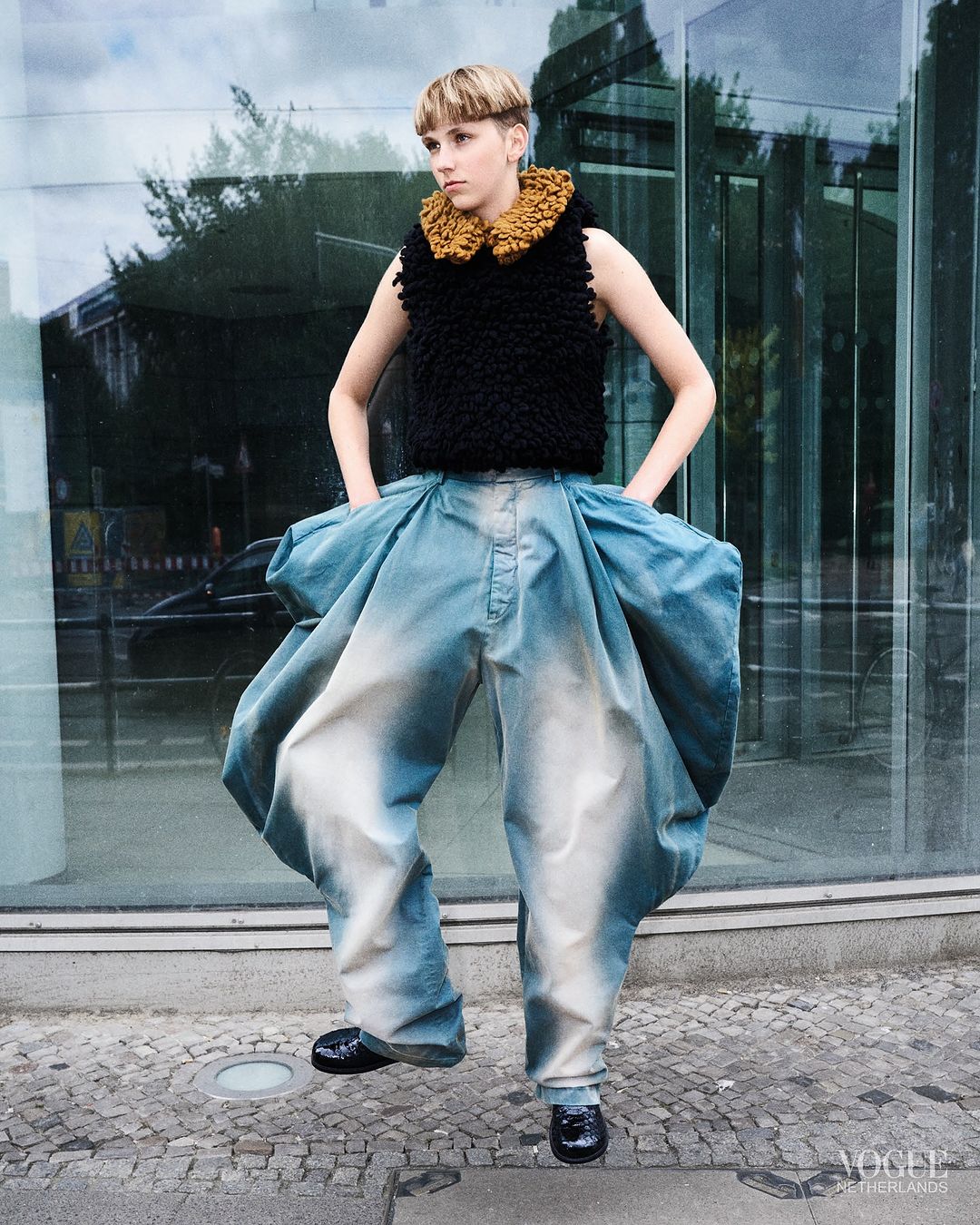 Person in avant-garde fashion poses outdoors. Dark top with a brown ruffled collar paired with light blue, voluminous pants and black shoes.