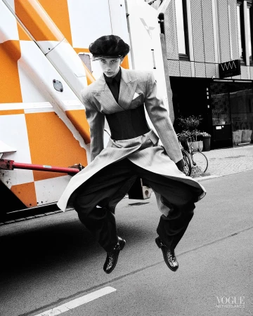 Model in motion mid-air wearing a tailored grey suit jacket, wide trousers, and a black cap on a Paris street.