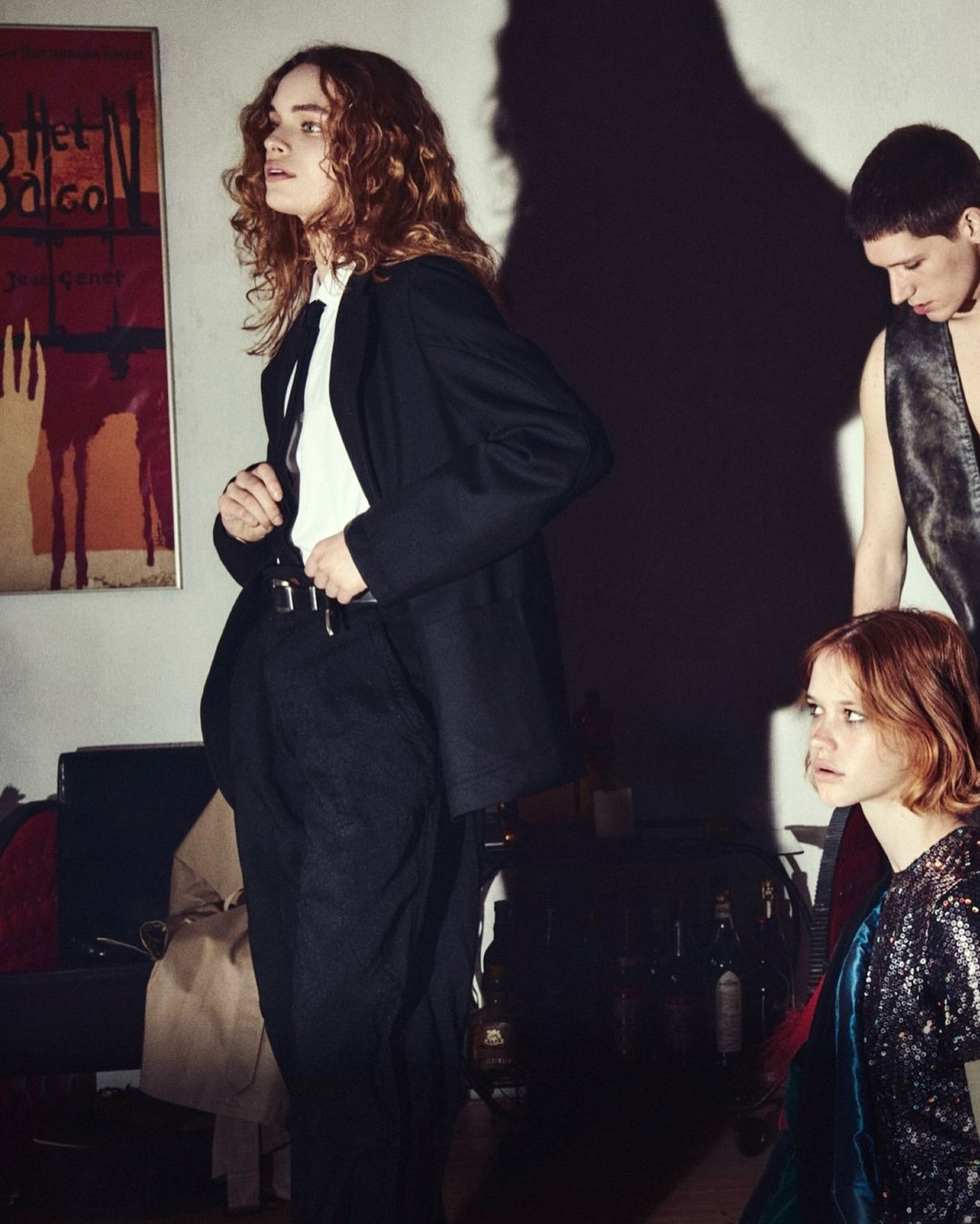 Group of three young adults in a dimly lit room with a vintage poster and dark furniture, evoking a casual, indoor scene.