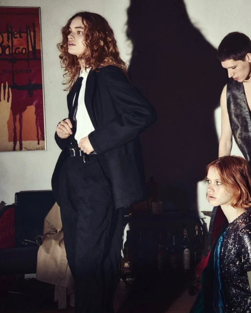 Group of three young adults in a dimly lit room with a vintage poster and dark furniture, evoking a casual, indoor scene.