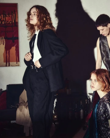 Group of three young adults in a dimly lit room with a vintage poster and dark furniture, evoking a casual, indoor scene.