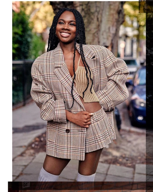 A smiling young woman with braids poses in a plaid blazer and pleated skirt on an outdoor sidewalk in an urban environment.