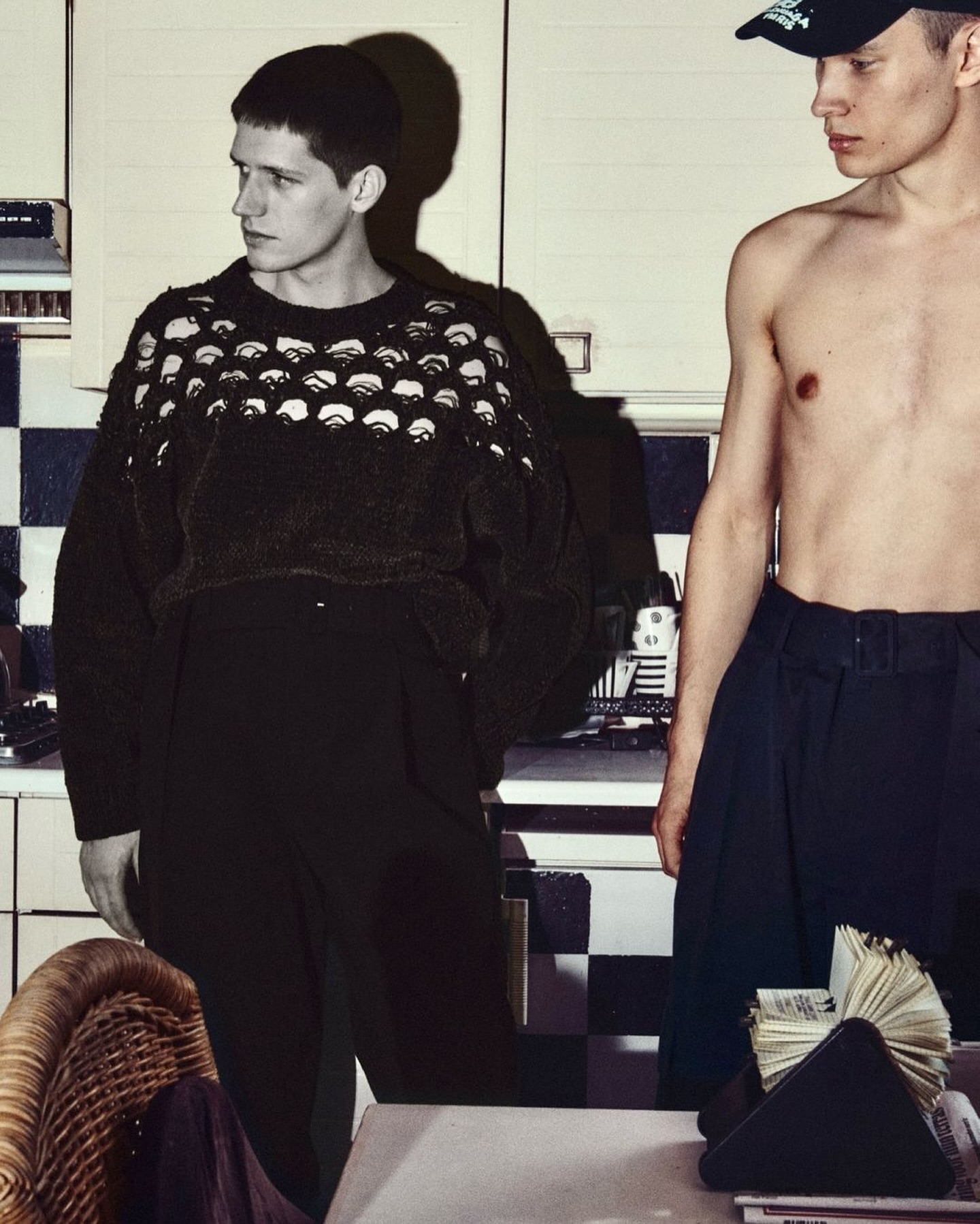 Two young adult men pose indoors; one wears a black patterned sweater and dark pants, while the other is shirtless with dark pants.