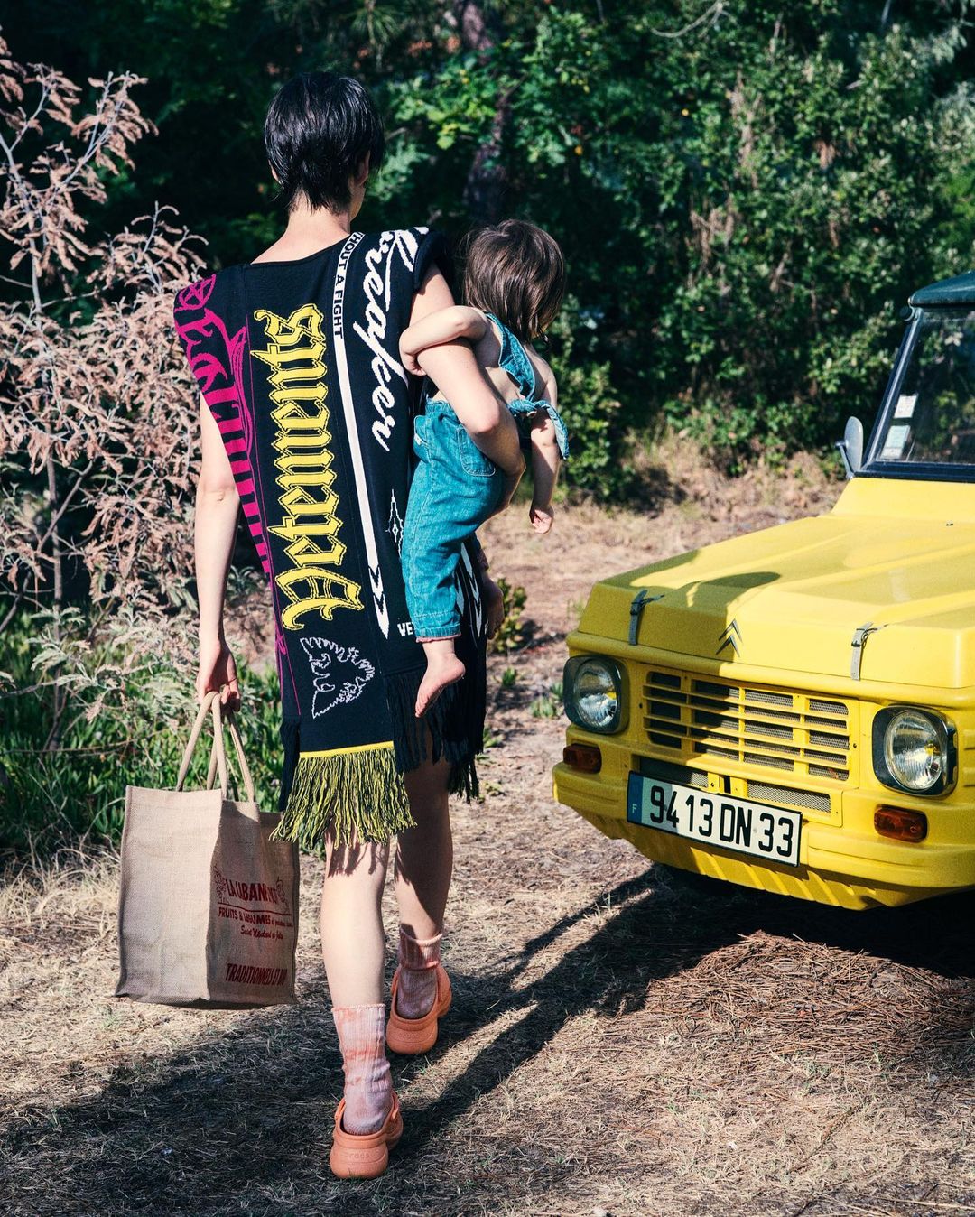 Woman carrying a child walking past a yellow car on a dirt path near foliage, wearing a black dress with fringe and text design.