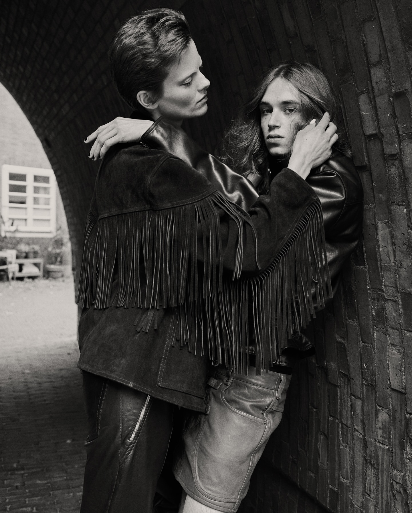 A black and white portrait of two adults with short hair, embracing in leather jackets in front of an archway in an urban setting.