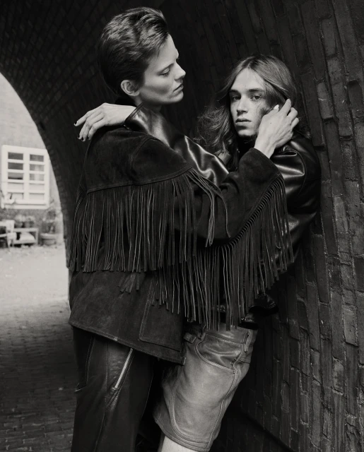 A black and white portrait of two adults with short hair, embracing in leather jackets in front of an archway in an urban setting.