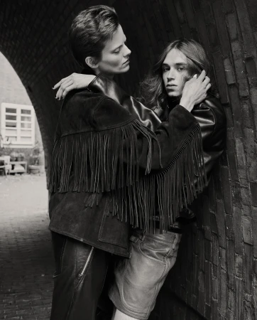 A black and white portrait of two adults with short hair, embracing in leather jackets in front of an archway in an urban setting.
