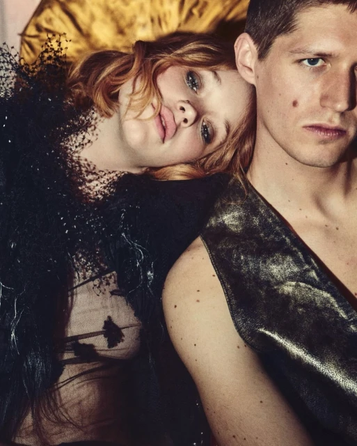 Fashionable young adult couple in an embrace: a woman with red hair and a black dress rests her head on a man's shoulder.