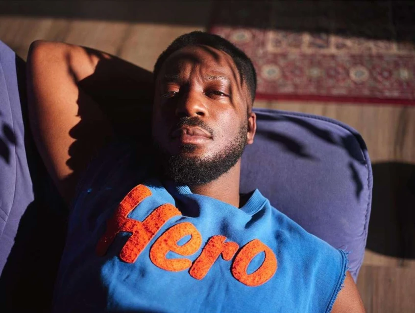 Adult man lies on a blue cushion, wearing a blue sleeveless shirt with the word Hero in orange lettering, bathed in sunlight.