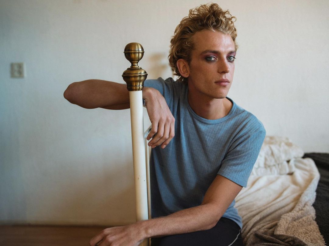 Portrait of a fair-skinned young adult male with blond curly hair and light makeup leaning on a white metal bed frame indoors.