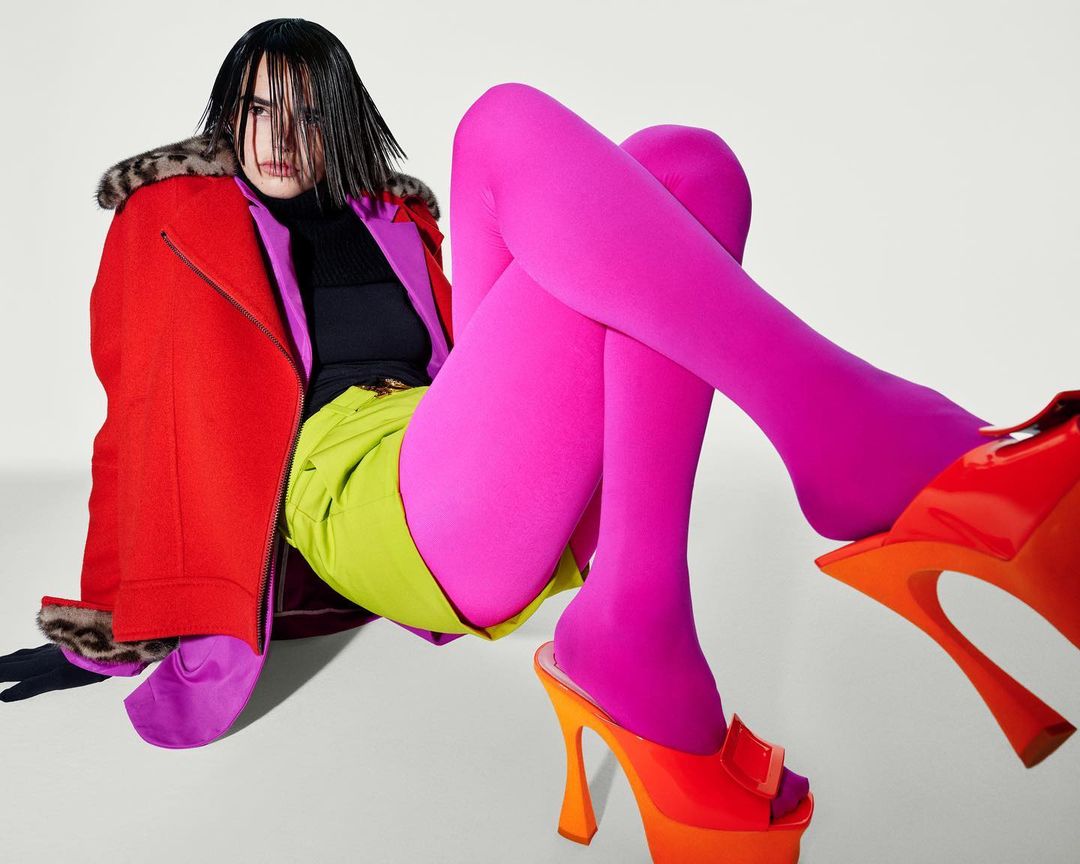 Fashion model poses in photo studio wearing a red jacket with fur trim, green shorts, hot pink tights and platform heels.