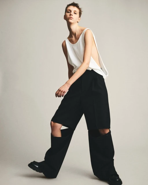 Fashion shoot of a slim adult woman with short hair wearing a white sleeveless top and baggy black pants with holes at the knees.
