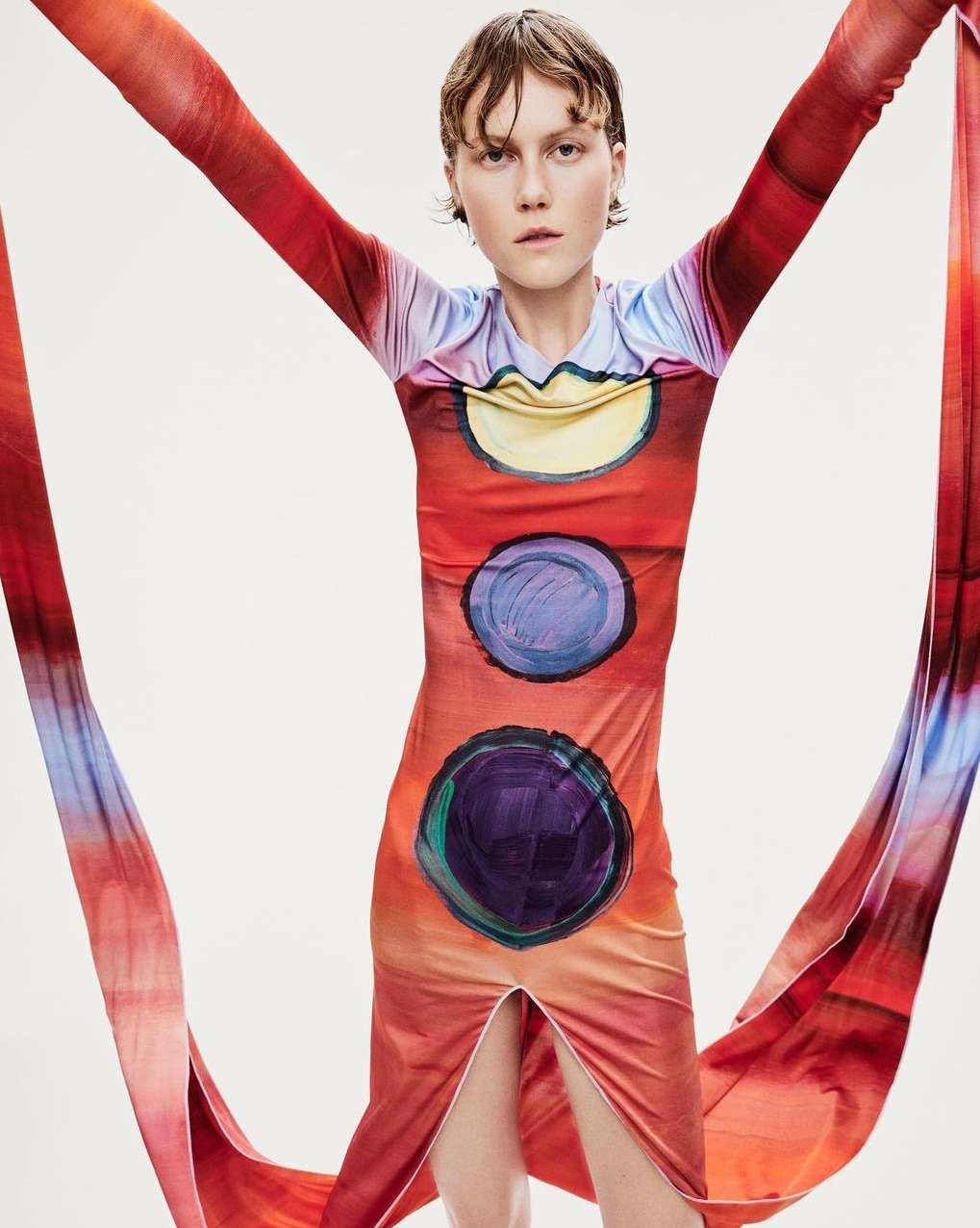 Adult woman in a colorful patterned dress with her arms up on a seamless white backdrop in a studio setting.