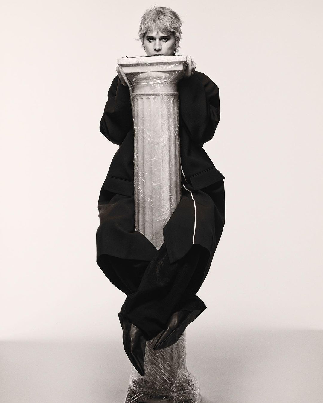 A person in a black suit sits atop a pillar wrapped in plastic in a monochromatic photo studio with a soft, diffused light, creating an editorial image.