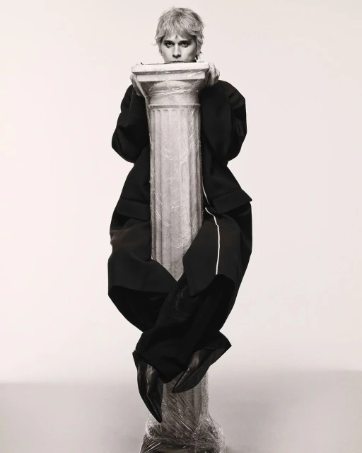 A person in a black suit sits atop a pillar wrapped in plastic in a monochromatic photo studio with a soft, diffused light, creating an editorial image.