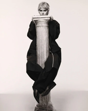 A person in a black suit sits atop a pillar wrapped in plastic in a monochromatic photo studio with a soft, diffused light, creating an editorial image.