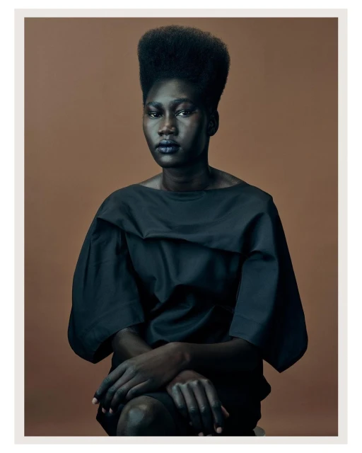 Striking portrait of a Black woman with a high hairstyle wearing a dark dress, seated against a neutral backdrop, in a studio setting.