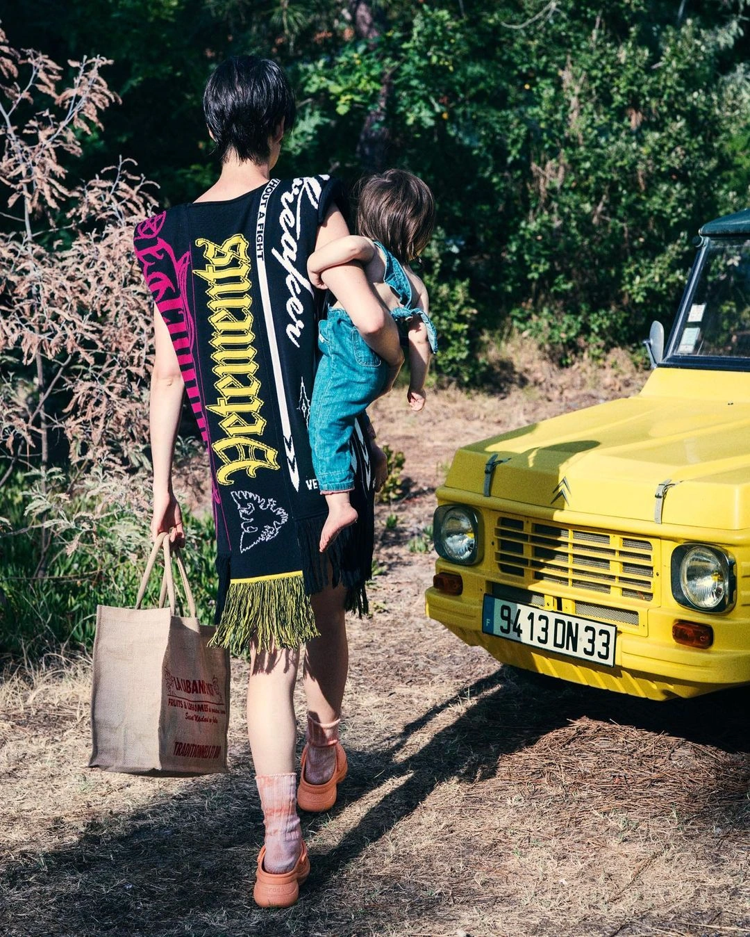 Woman carrying a young child while walking past a yellow car on a dirt path, holding a woven bag.