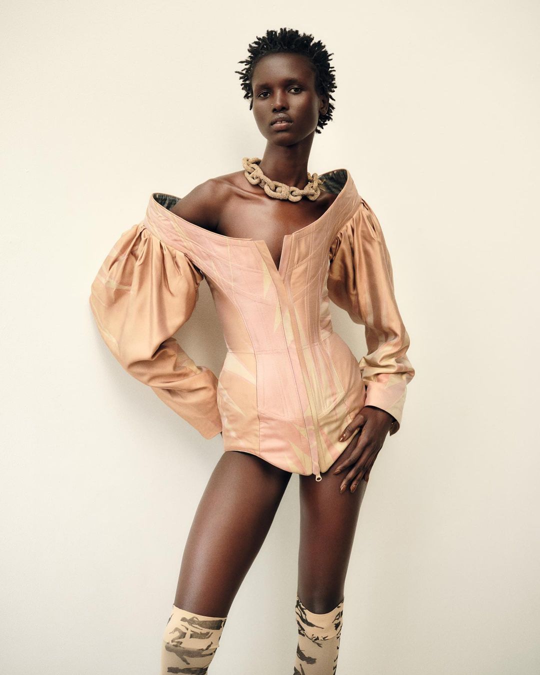 Fashion portrait of a Black woman wearing a pink couture corset top and patterned thigh-high boots against a light neutral background.