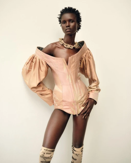 Fashion portrait of a Black woman wearing a pink couture corset top and patterned thigh-high boots against a light neutral background.
