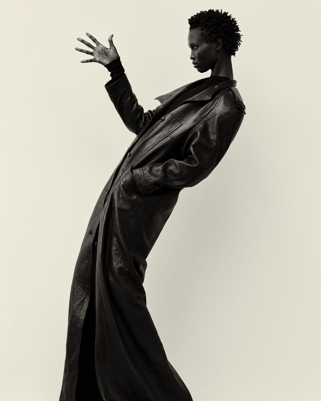 Dramatic black-and-white portrait of a Black female model in a long leather coat striking a pose in a photo studio setting.