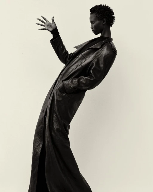 Dramatic black-and-white portrait of a Black female model in a long leather coat striking a pose in a photo studio setting.