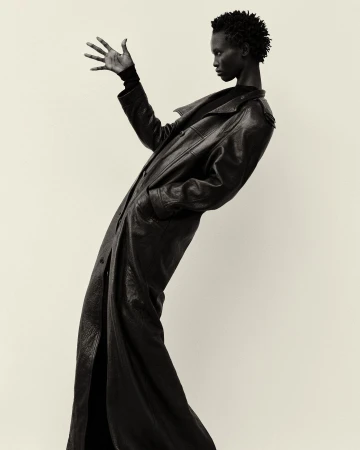 Dramatic black-and-white portrait of a Black female model in a long leather coat striking a pose in a photo studio setting.