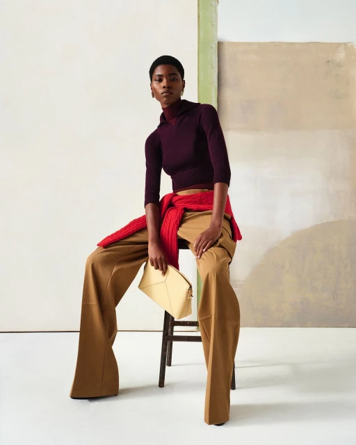 Fashionable woman in burgundy top and wide-leg trousers, seated, holding beige clutch, with a red sweater around her shoulders.