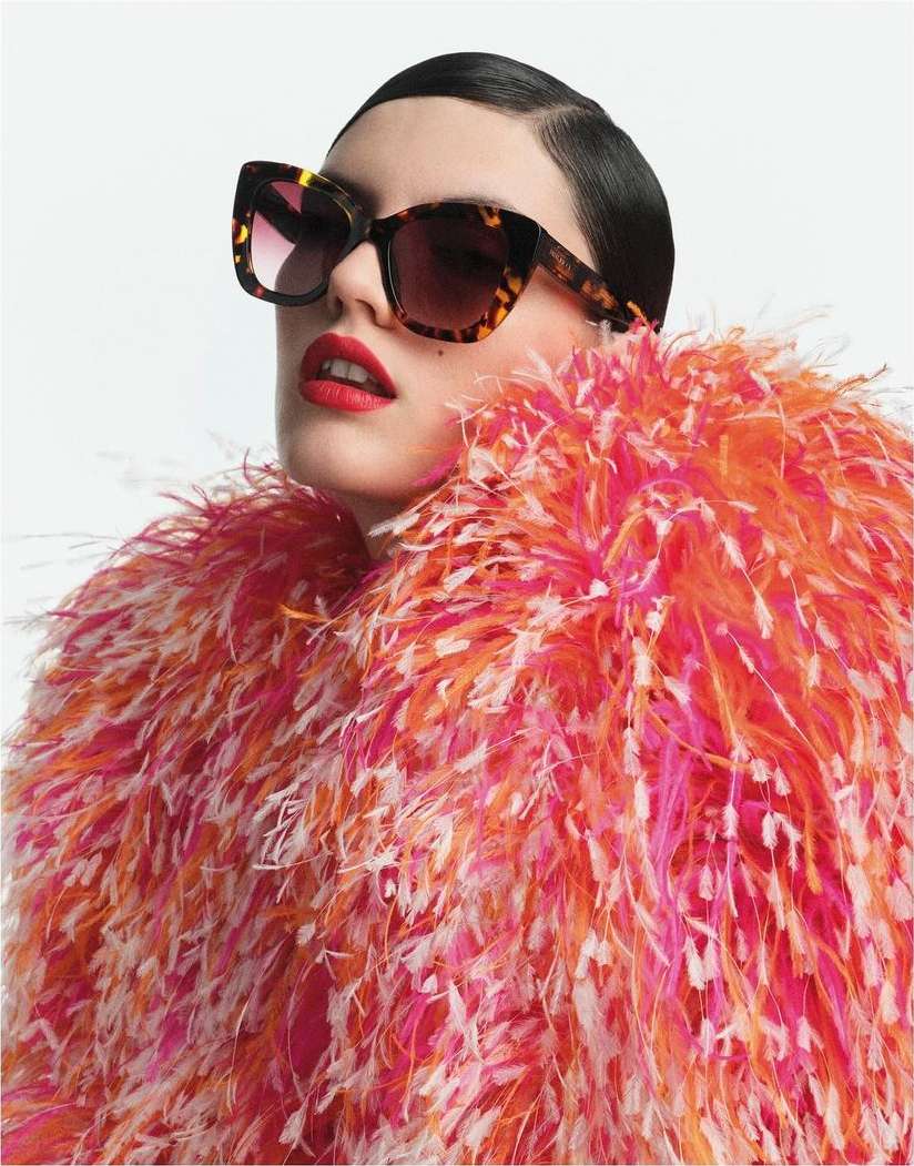 Woman wearing tortoiseshell sunglasses and a vibrant pink and orange feather coat against a white background