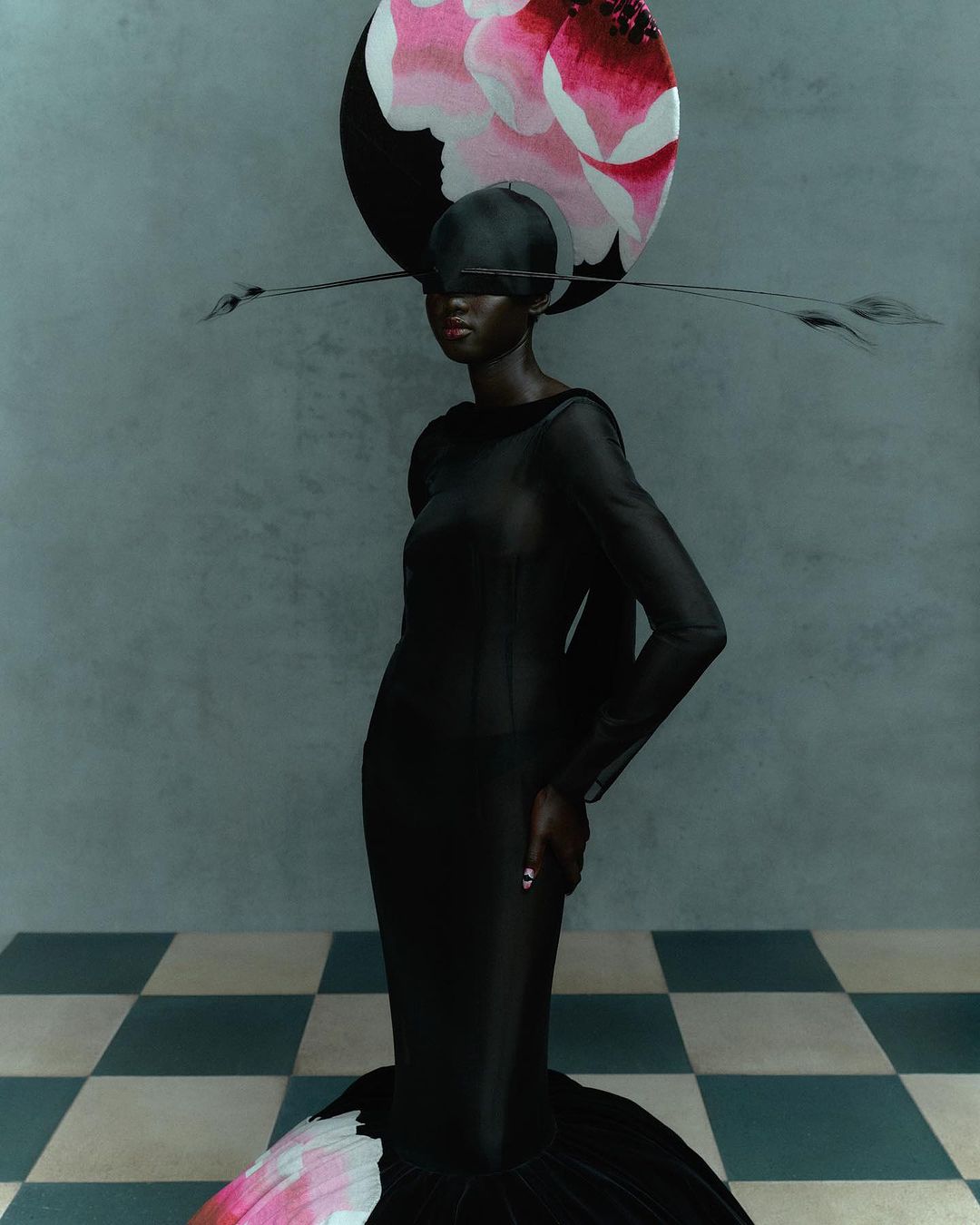 Fashionable African woman wearing a black gown and large floral headpiece poses in a muted-tone studio with checkerboard flooring.